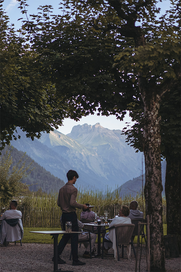 Gedeckte Tische unter Bäumen auf der Terrasse, dahinter die Allgäuer Berge